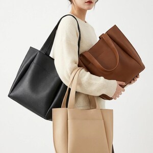 May include: Three leather tote bags in black, tan, and brown. Each bag has a rectangular shape with a top handle and a front pocket. The bags are held by a person wearing a cream-coloured jumper.