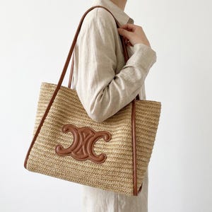 May include: A woven straw tote bag with brown leather trim and handles. The bag features a brown leather emblem on the front. The bag is rectangular and appears to be a summer accessory.