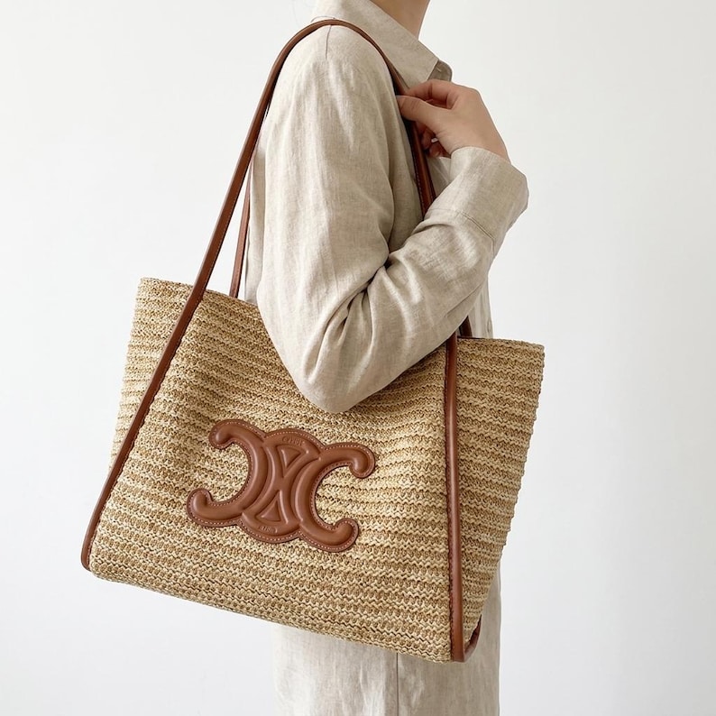 May include: A woven straw tote bag with brown leather trim and handles. The bag features a brown leather emblem on the front. The bag is rectangular and appears to be a summer accessory.
