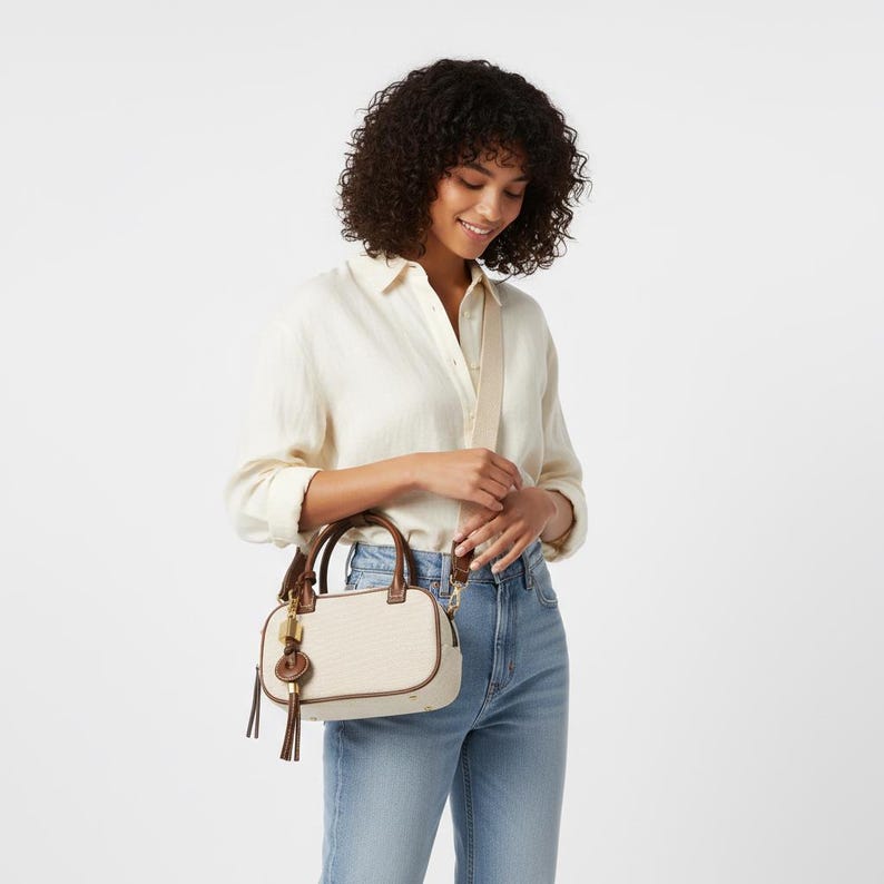 May include: A cream-coloured handbag with brown leather trim and handles. The bag has a detachable shoulder strap and a decorative tassel. The bag is rectangular and appears to be made of canvas material.