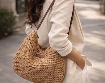 May include: A woven straw shoulder bag with brown leather straps and a small brown leather tag. The bag has a rounded shape and a textured surface, perfect for carrying essentials.