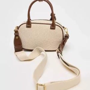 May include: Cream-coloured handbag with brown leather accents. The bag features a top handle and a detachable shoulder strap. The bag's exterior is a woven fabric, and the trim and handle are brown leather. The strap is cream-coloured with gold-coloured hardware.