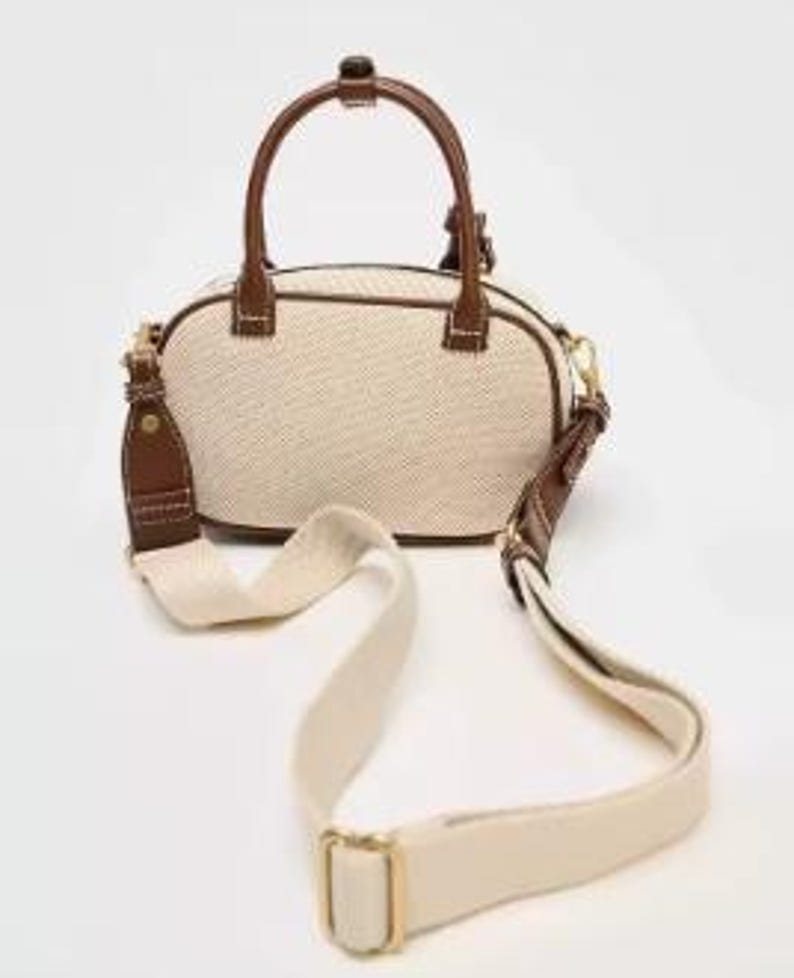 May include: Cream-coloured handbag with brown leather accents. The bag features a top handle and a detachable shoulder strap. The bag's exterior is a woven fabric, and the trim and handle are brown leather. The strap is cream-coloured with gold-coloured hardware.