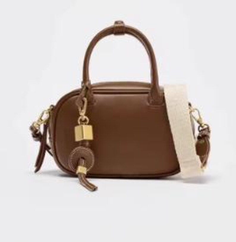 May include: A brown leather handbag with a top handle and a detachable cream-coloured shoulder strap. The bag features gold-toned hardware, including a decorative charm with a square and circular element. The bag is oval-shaped and sits on a white surface.