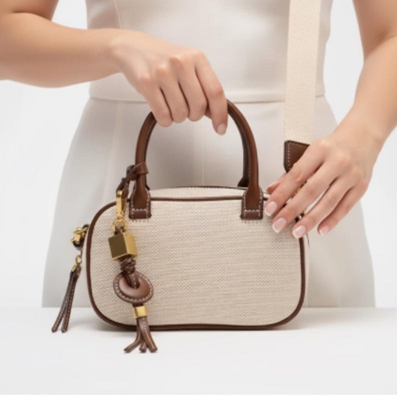 May include: A beige handbag with brown leather handles and trim. The bag has a rectangular shape with rounded corners and a detachable shoulder strap. A gold-coloured charm with a key and tassel hangs from the side.