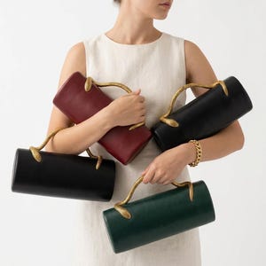 Op de afbeelding: Vier cilindrische handtassen in bordeaux, zwart en groen, elk met een gouden slangvormige handgreep. De tassen worden vastgehouden door een persoon in een witte jurk. Een gouden armband is zichtbaar.