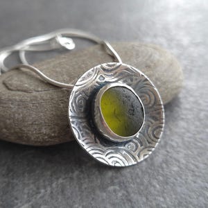 May include: A silver pendant necklace featuring a circular, textured pendant with a central bezel-set, olive-green sea glass stone. The pendant hangs from a delicate silver chain, showcasing a unique, handcrafted design.