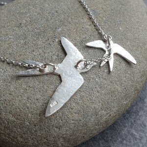 Peut inclure: Collier en argent avec deux d&eacute;licats pendentifs en forme d'oiseau en argent. Les oiseaux sont orient&eacute;s dans la m&ecirc;me direction et sont reli&eacute;s par une petite cha&icirc;ne. Le collier est pr&eacute;sent&eacute; sur un fond de pierre grise.