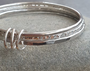 May include: Close-up of a silver bangle bracelet. The bracelet features three bands, with one band having a textured, hammered design. The bands are connected with coiled silver wire. The bracelet is displayed on a gray surface.