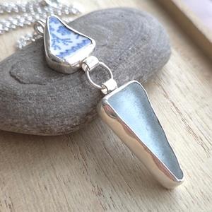 May include: A silver necklace featuring two pendants. One pendant is a blue and white patterned ceramic piece, and the other is a light blue, sea glass-like shape. The pendants are connected by a silver ring and chain.