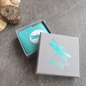 Op de afbeelding: Een grijze juwelendoos met een turquoise libellenontwerp en de tekst "dragonfly Lane". De doos is open en bekleed met turquoise tissuepapier.