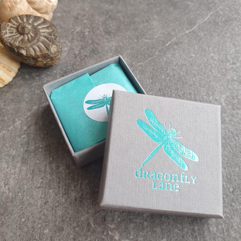 Op de afbeelding: Een grijze juwelendoos met een turquoise libellenontwerp en de tekst "dragonfly Lane". De doos is open en bekleed met turquoise tissuepapier.