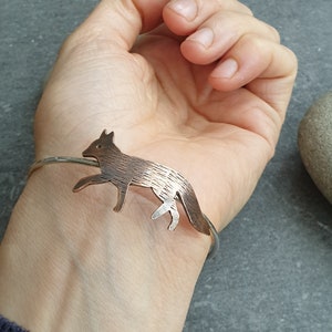May include: A silver bangle bracelet with a copper fox charm. The fox is detailed with textured fur and is in a running pose.
