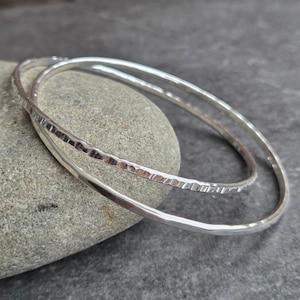 Peut inclure: Deux bracelets argentés, dont un avec une texture martelée, reposent sur une pierre grise sur fond sombre. Les bracelets circulaires reflètent la lumière, présentant un design de bijoux minimaliste.