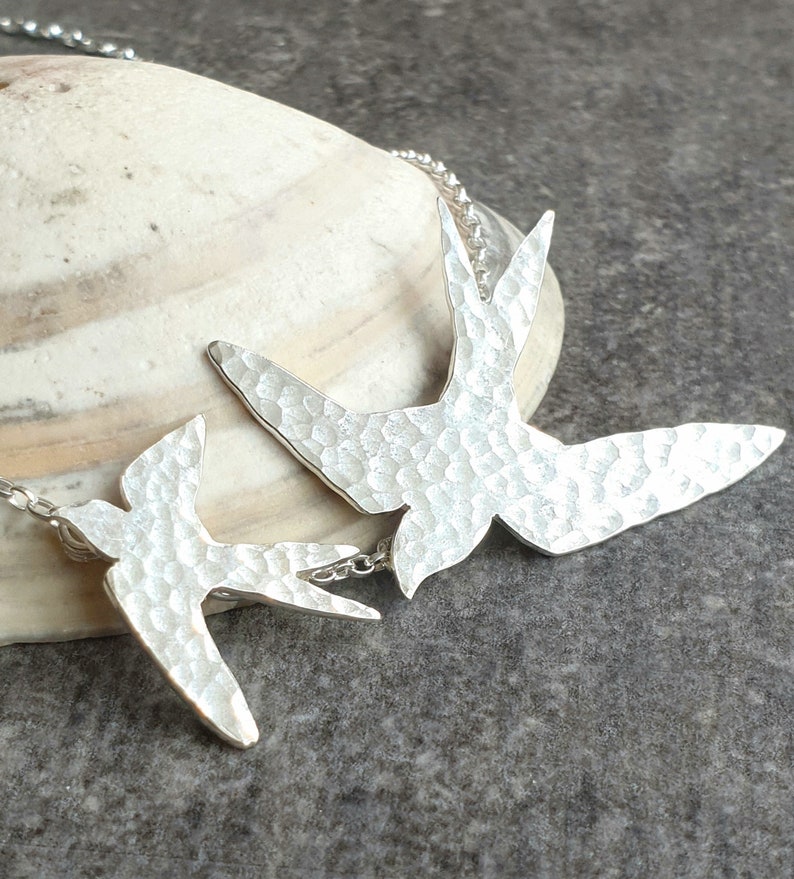 Peut inclure: Un collier en argent avec deux pendentifs en forme d'hirondelle en argent martel&eacute;. Les oiseaux sont orient&eacute;s dans la m&ecirc;me direction et sont reli&eacute;s par une petite cha&icirc;ne.