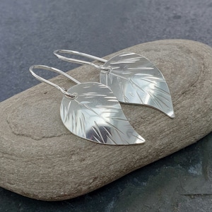 May include: A pair of silver leaf earrings with a textured design. The earrings are hanging from a silver hook.