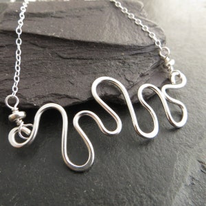 Op de afbeelding: Zilveren ketting met een golvend, abstract ontwerp. De ketting is gemaakt van een enkel stuk draad dat is gebogen in een reeks bochten.