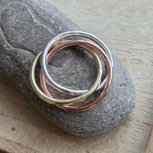May include: A set of four interlocking rings in gold, silver, and rose gold tones. The rings have a hammered texture and are arranged on a gray stone surface. The rings are a jewelry item.