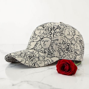 May include: A cream-coloured baseball cap with a repeating pattern of abstract faces in black. The cap has a curved brim and a structured crown. A red rose lies next to the cap on a white surface.