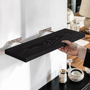 May include: A black wooden shelf with a natural wood grain pattern is held up by two silver metal brackets. The shelf is rectangular and appears to be about 30 inches long. The shelf is mounted on a white wall.