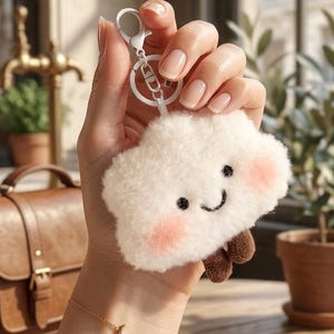 May include: A fluffy white cloud keychain with a smiling face, rosy cheeks, and brown feet. The keychain has a silver clasp. The background includes a leather bag, plants, and a wooden table.