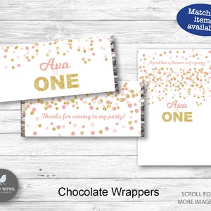 May include: Pink and gold confetti chocolate bar wrappers with the text "Ava is ONE" printed on them. The wrappers are designed for a first birthday party.