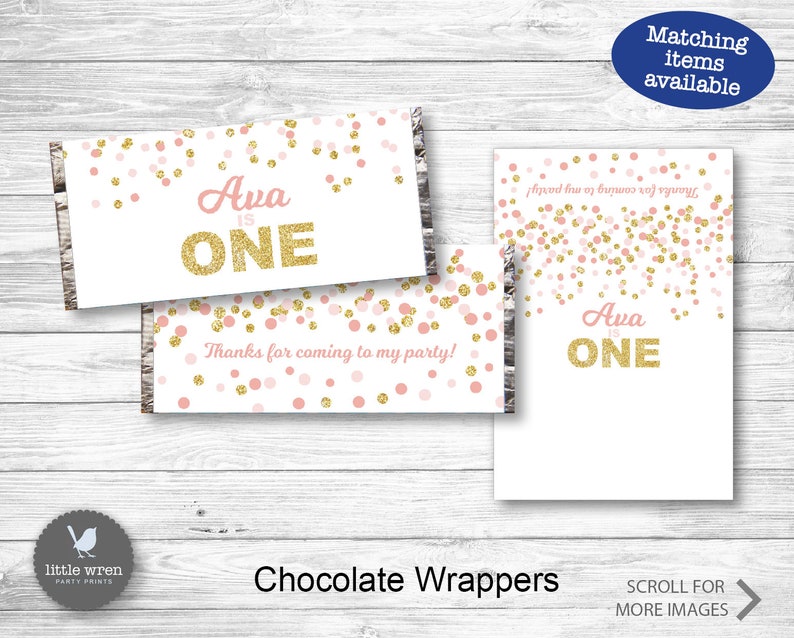 May include: Pink and gold confetti chocolate bar wrappers with the text "Ava is ONE" printed on them. The wrappers are designed for a first birthday party.