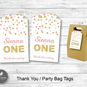 May include: Two pink and gold thank you tags with the text "Sienna is One" and "Thanks for coming". The tags are decorated with pink and gold confetti. A brown paper bag with a handle is also shown with a smaller version of the tag attached.