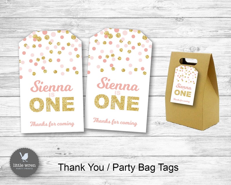 May include: Two pink and gold thank you tags with the text "Sienna is One" and "Thanks for coming". The tags are decorated with pink and gold confetti. A brown paper bag with a handle is also shown with a smaller version of the tag attached.
