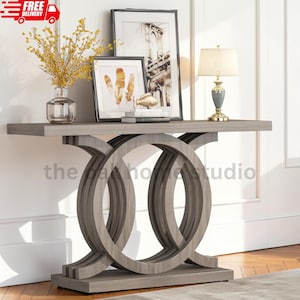 May include: A gray wood console table featuring a rectangular top and a base with two large interlocking circular supports. On the table sits a clear glass vase with yellow flowers, two framed prints, a small gold fish figurine, and a table lamp.