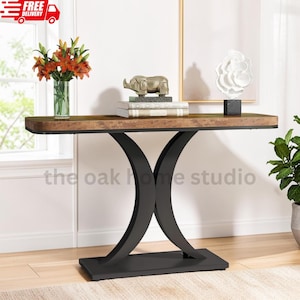 May include: A console table with a rectangular wood-grain top and a black metal base featuring two curved, intersecting legs. The table holds a glass vase with orange lilies, a stack of books topped with a gold rhino figurine, and a white sculpture.