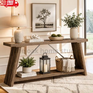 May include: A long wooden console table with an X-frame base and a lower shelf. The table holds a ceramic lamp, a decorative knot, books, a bowl of moss balls, a plant, a black metal lantern, and a woven basket with a white fringed throw blanket.