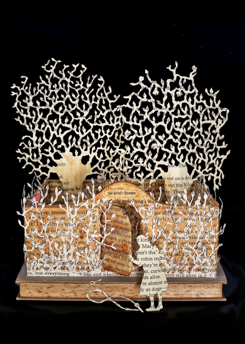 May include: A papercraft sculpture of a girl standing in front of a book with the title "The Secret Garden" on the cover. The book is open and the pages are cut out to create a garden scene with trees and flowers. The girl is holding a book in her hand.