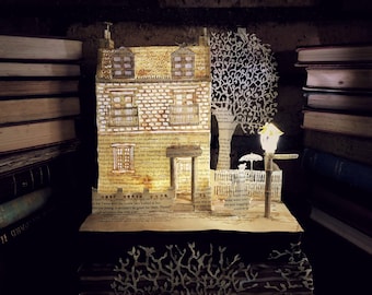 May include: A miniature paper house with a lit lamppost and a tree made of paper, set against a backdrop of stacked books. The house is made of pages from a book, with the text visible. The house has a door with the words "Mary Poppins" above it.