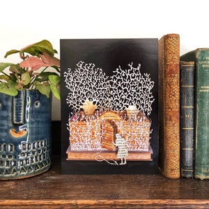 May include: A black card with a white paper cut-out design of a forest scene. The forest is made of paper and has a white, paper cut-out figure of a person standing in the middle of the scene. The card is on a wooden shelf with a potted plant and a row of books behind it.