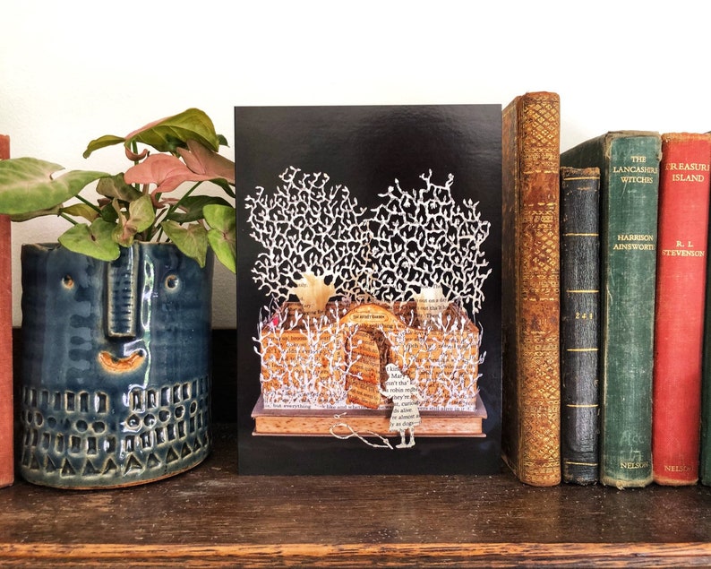 May include: A black card with a white paper cut-out design of a forest scene. The forest is made of paper and has a white, paper cut-out figure of a person standing in the middle of the scene. The card is on a wooden shelf with a potted plant and a row of books behind it.