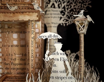 May include: A papercraft diorama of Mary Poppins walking with an umbrella in a garden setting. The scene is made from pages of a book, with the text visible. The diorama includes a lamppost, a tree, and a house.