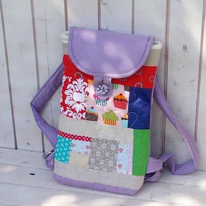 Può includere: Uno zaino in patchwork viola con chiusura a lembo e bottone. Lo zaino è realizzato con diversi tessuti con vari motivi, tra cui cupcake, fiori e pois.