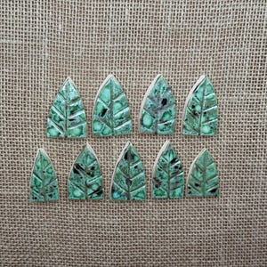 May include: Ten ceramic leaf-shaped tiles with a glossy, mottled green glaze. The tiles have a textured surface with visible veins and are arranged on a textured, tan fabric background. The leaves are approximately 1.5 inches tall.