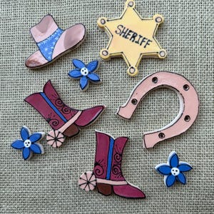 May include: A collection of ceramic western-themed embellishments including a yellow sheriff star badge, two pink and blue cowboy boots with spurs, a pink horseshoe, a pink cowboy hat with a blue band, and three small blue five-petaled flowers.