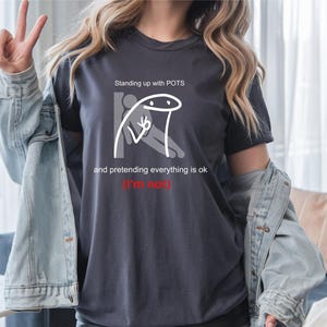 May include: Dark gray t-shirt with the text "Standing up with POTS and pretending everything is ok (I'm not)" and a stick figure graphic. The shirt is a crew neck and has short sleeves.