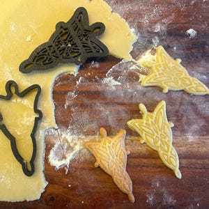 Puede incluir: Primer plano de cortadores de galletas y galletas en forma de hoja estilizada. Un cortador de galletas negro está presionado en la masa, y tres galletas están sobre una superficie de madera. Las galletas son de color amarillo claro y tienen diseños intrincados.