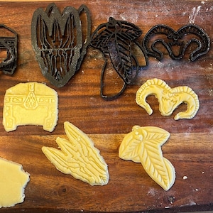Pode incluir: Uma coleção de cortadores de biscoitos de plástico preto e massa cortada em uma superfície de madeira. Os cortadores têm várias formas, incluindo um capacete, um baú e uma folha. A massa é amarelo claro.