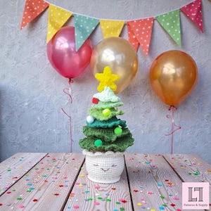 Pode incluir: Uma árvore de Natal de crochê feita à mão em um vaso branco com um rosto sorridente. A árvore é em vários tons de verde, coberta com uma estrela amarela e decorada com pompons coloridos. Balões e uma faixa no fundo.