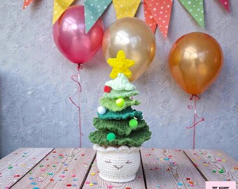 Könnte beinhalten: Ein handgehäkelter Weihnachtsbaum in einem weißen Topf mit einem lächelnden Gesicht. Der Baum ist in verschiedenen Grüntönen gehalten, mit einem gelben Stern gekrönt und mit bunten Pompons verziert. Luftballons und ein Banner sind im Hintergrund.