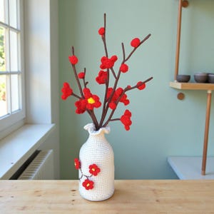 Puede incluir: Un jarrón blanco de ganchillo con flores rojas de ganchillo y tallos marrones. El jarrón tiene una parte superior con volantes y está decorado con flores rojas adicionales. Las flores tienen centros amarillos. El jarrón está sobre una superficie de madera.