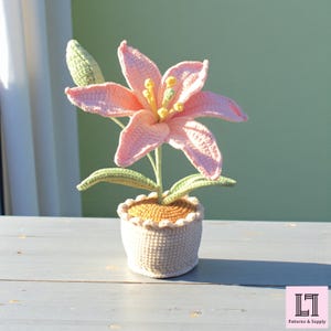 May include: A crocheted lily flower in a white pot. The flower has pink petals, yellow stamens, and green leaves. The pot is white with a scalloped edge and sits on a gray surface. The overall theme is home decor.