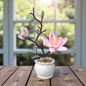 May include: A handcrafted crochet flower arrangement in a white pot. The arrangement features pink flowers with yellow centers, green leaves, and brown stems. The pot has a scalloped edge and sits on a wooden surface.