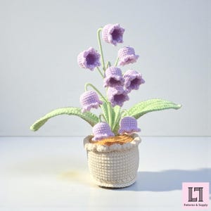May include: A crocheted lily of the valley plant in a cream-colored pot. The plant features light purple bell-shaped flowers, green leaves, and a light brown base. The pot has a scalloped edge. The image includes the text "Patterns & Supply".