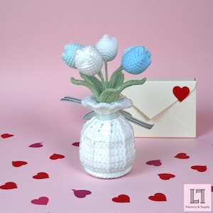 May include: A handmade crochet bouquet of tulips in a white vase, with blue and white flower heads and green leaves. A cream-colored envelope with a red heart is in the background, along with scattered heart-shaped confetti. The vase is tied with a light blue ribbon.